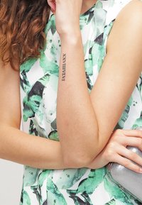 Zielona i biała bluzka w wzory z abstrakcyjnymi motywami; srebrna kopertówka trzymana w jednej ręce; widoczny tatuaż na nadgarstku.