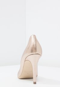 Escarpin à talon haut en métal doré rose, vu de derrière sur une surface blanche avec un fond blanc uni.