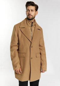 Palton din amestec de lână de camel cu guler cu fante, două buzunare frontale și butoane vizibile. Purtați peste o jachetă puf cu fermoar. Pantaloni întunecați dedesubt.