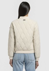 Ljusbeige quiltad jacka med en figursydd design, ribbad muddar och nederkant samt en liten baketikett. Slät textur med diamantmönstrad sömnad.