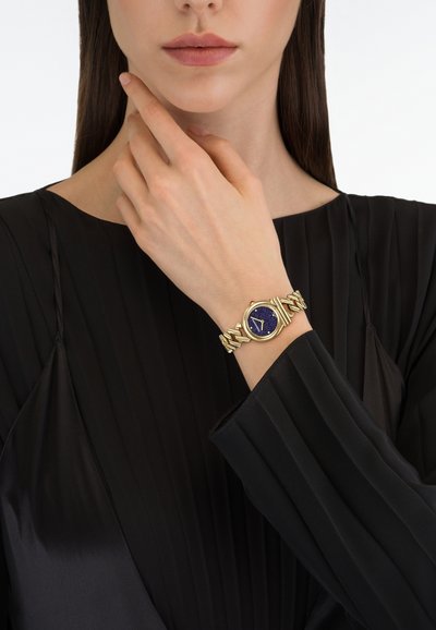 Zlaté hodinky s modrým texturovaným ciferníkem a svítícími ručičkami. Náramek z řetízkových článků. Na ruce s neutrální barvou nehtů. Černý plisovaný top.