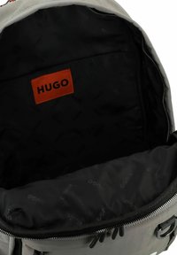 Zaino grigio con apertura con zip. L'interno presenta una fodera nera, il marchio "HUGO" e una trama di tessuto liscia. Nessuna tasca visibile.