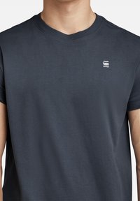 Grå bomullströja med rundad halsringning och korta ärmar. Har en liten vit logotyp på vänster bröstområde. Slät textur.