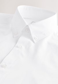 Chemise blanche à boutons avec col pointu et boutons visibles, étalée à plat sur une surface claire.