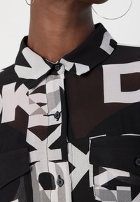Gros plan d'une chemise boutonnée à motif noir et blanc avec un col et des poches à rabat, portée par une personne avec des boucles d'oreilles créoles.