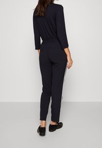 Marineblauwe jumpsuit met lange mouwen, een elastische tailleband en witte zijkanten. Zacht stof, tapered benen en gecombineerd met zwarte schoenen.