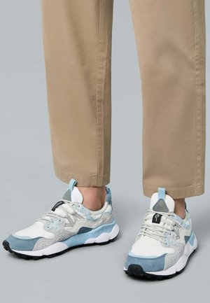 Zapatillas deportivas con una mezcla de materiales de ante y malla en blanco, azul claro y gris; con una suela texturizada y un sistema de cordones destacado.