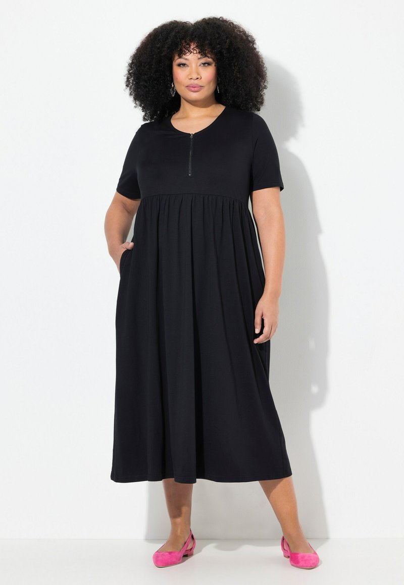 Vestido negro con mangas cortas y un escote con cremallera, que presenta una cintura fruncida y bolsillos, combinado con unos slippers rosas. Textura de tela suave.