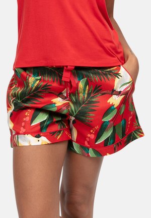 Kobieta nosząca czerwone spodenki w tropikalny wzór z zielonymi liśćmi i białymi ptakami, w połączeniu z czerwoną koszulą, ręka w kieszeni.
