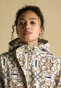 Jauna sieviete ar cirtainiem matiem, valkājot zelta auskarus gredzenu veidā un baltu jaku ar elementiem, kas izskatās pēc virvēm, sprādziņām un jostām, skatās uz priekšu uz bēša fona.