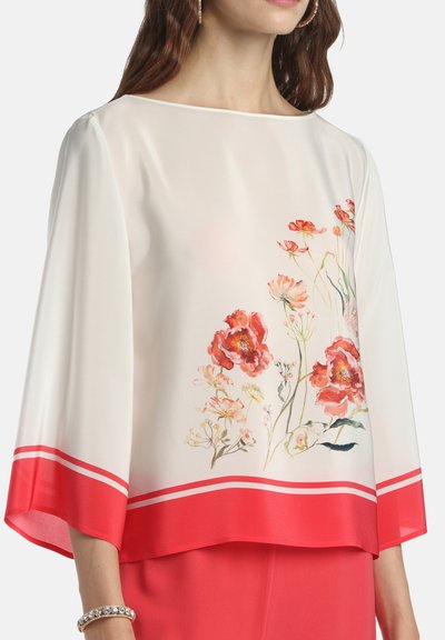 Blouse blanche à manches larges, imprimé floral rouge sur le devant, et bordure rouge à l'ourlet, portée avec une jupe rouge assortie.