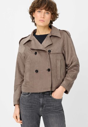 Frau mit kurzen lockigen Haaren trägt eine zweireihige Jacke in Taupe über einem schwarzen Pullover und dunkelgraue Jeans, eine Hand in der Tasche.