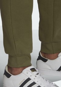 Olivgröna mjukisbyxor med ribbade muddar, kombinerade med vita sneakers med svarta detaljer och gyllene märkning.