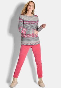Grauer Strickpullover mit pinken geometrischen Mustern, kombiniert mit schmal geschnittenen rosa Cordhosen. Das Modell trägt hellbeige schnürbare Stiefel.