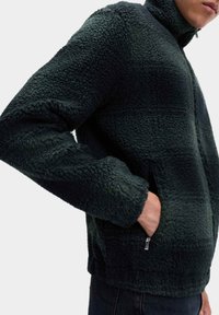 Ciemnozielona polarowa kurtka z wysokim kołnierzem, teksturowaną powierzchnią i zapinanymi na zamek bocznymi kieszeniami. Charakteryzuje się luźnym krojem i miękkim materiałem.
