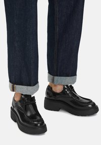 Persoană îmbrăcată cu blugi negri răsuciți și încălțăminte neagră lucioasă cu toc chunky și șireturi pe un fundal deschis.