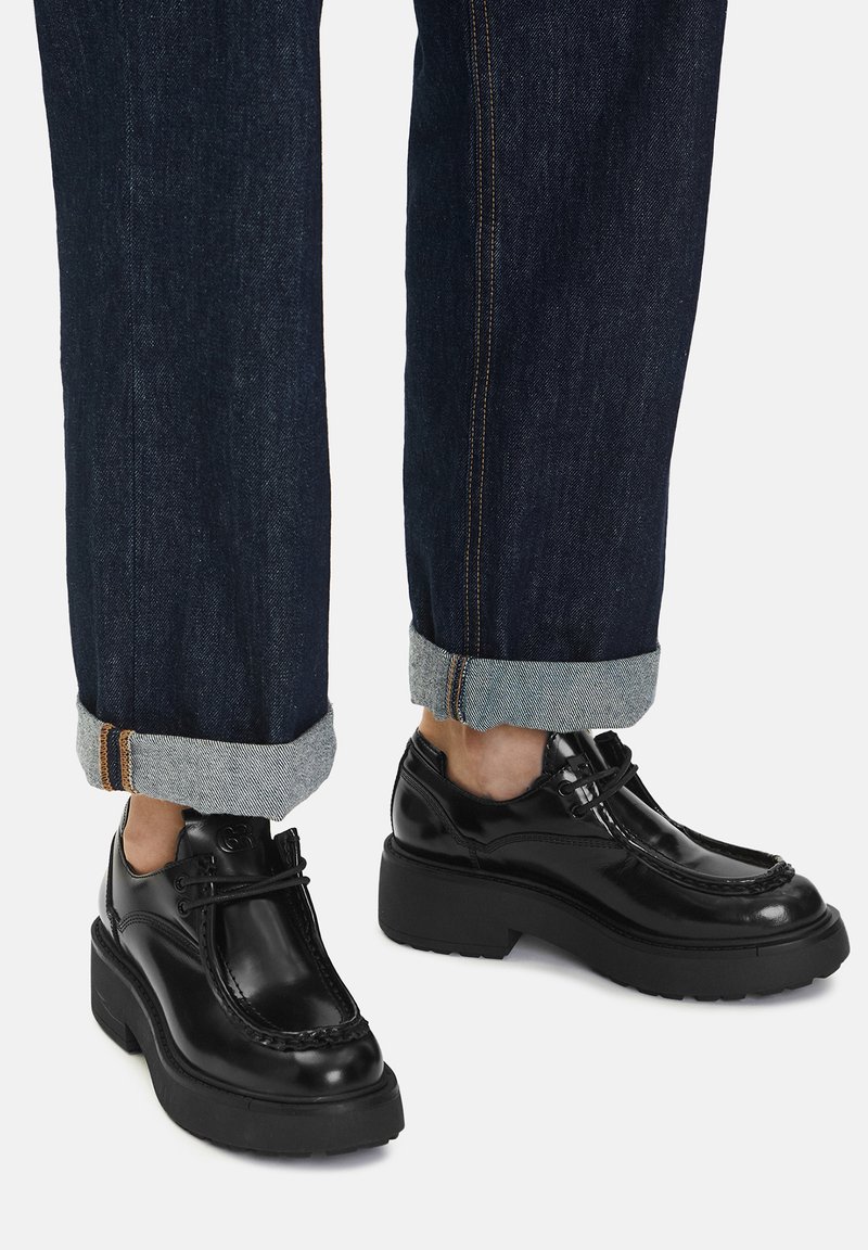 Persoană îmbrăcată cu blugi negri răsuciți și încălțăminte neagră lucioasă cu toc chunky și șireturi pe un fundal deschis.