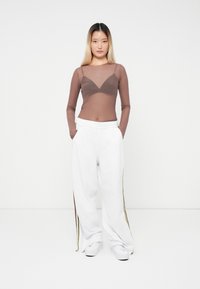 Brown mesh long-sleeve top paired with white, wide-leg pants featuring side stripes in green and burgundy. Model standing with hands in pockets.