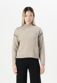 Donna con capelli lunghi e lisci che indossa un maglione beige a collo alto con dettagli di cucitura nera e pantaloni neri, in piedi di fronte a uno sfondo bianco.