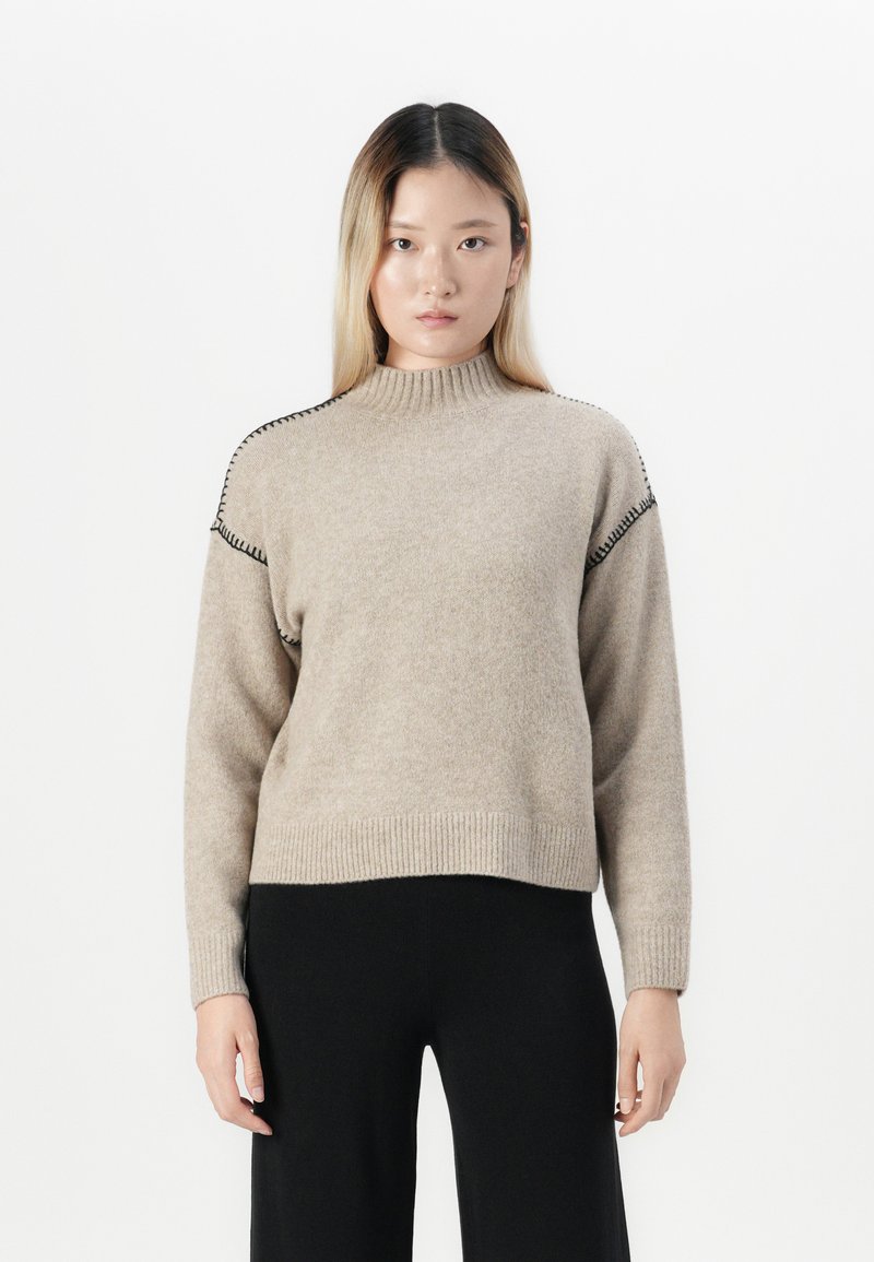 Donna con capelli lunghi e lisci che indossa un maglione beige a collo alto con dettagli di cucitura nera e pantaloni neri, in piedi di fronte a uno sfondo bianco.