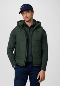 Gilet imbottito verde scuro con zip, dotato di cappuccio e logo a diamante. Indossato sopra una maglia scura, abbinato a pantaloni blu navy.