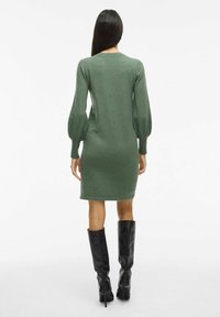 Grünes gestricktes Kleid mit langen Puffärmel, eng anliegendem Oberteil und knielangem Saum. Tragen mit hohen schwarzen Stiefeln. Strukturierter Stoff.