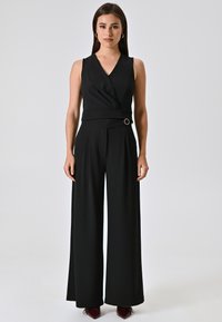 Zwarte jumpsuit met een V-hals, wikkelontwerp, wijde pijpen en een riem met een cirkelvormige accenten. Glad stof met een gestructureerde uitstraling.