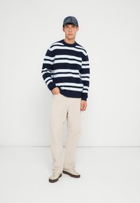 Maglione a righe navy e crema con collo a giro, abbinato a pantaloni beige e sneakers chiare. Il modello indossa un cappellino blu scuro.