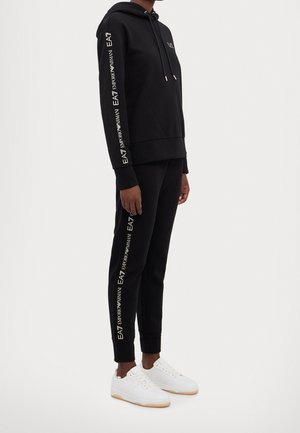 Persoon draagt zwarte capuchontrui en bijpassende joggers met witte EA7 Emporio Armani-logo's op de mouwen en broekspijpen, gecombineerd met witte sneakers.