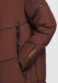 Jachetă puffer maro cu textură matlasată, având un fermoar frontal și două buzunare laterale. Unul dintre mâneci are un buzunar cu fermoar dotat cu accesorii negre.