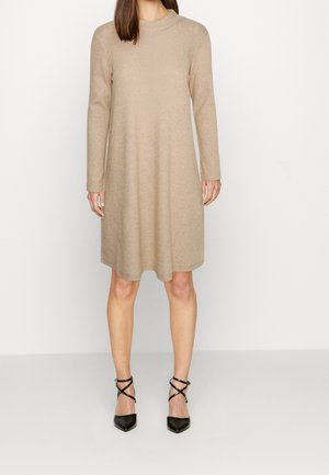 Kobieta w beżowej, dzianinowej sukience z długim rękawem do kolan, w połączeniu z czarnymi szpilkami na kostkę z paskiem, stojąca na białej podłodze.