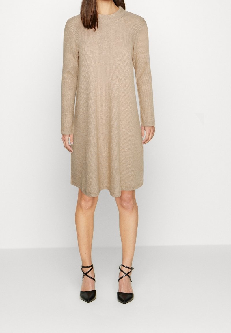 Femme portant une robe en maille beige à manches longues et longueur genou, associée à des escarpins noirs à bride cheville et bout pointu, debout sur un sol blanc.