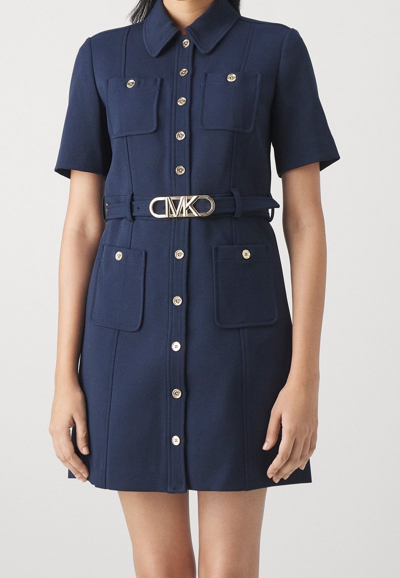 Robe bleu marine à manches courtes avec col, fermeture à boutons sur le devant, deux poches poitrine et deux poches hanches, et ceinture avec boucle logo MK dorée.