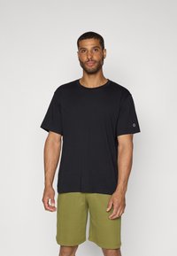 Svart bomulls t-skjorte med kort erme, rund hals, med en liten logo på venstre erme, kombinert med olivengrønne shorts.