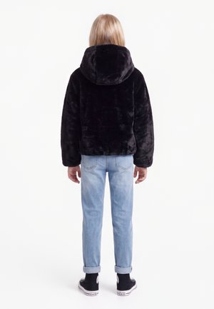Enfant portant une veste noire à capuche duveteuse, un jean bleu clair retroussé et des baskets montantes noires, debout de dos sur un fond blanc.