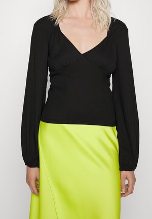 Zwarte langdraadige top met een diepe V-hals en geplooide details. Gecombineerd met een zijden limoen groene rok met een diagonale naad.