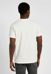 Camiseta corta de manga corta color crema, hecha de algodón, con cuello redondo, corte relajado y textura suave, vista desde la parte de atrás.