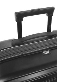 Maleta rígida negra con asa retráctil, apertura con cremallera y etiqueta de logo. Superficie suave y diseño minimalista.
