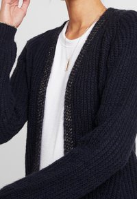 Cardigan lavorato a maglia di colore blu navy con una finitura testurizzata e apertura frontale, caratterizzato da un sottile dettaglio di filo metallico lungo i bordi.