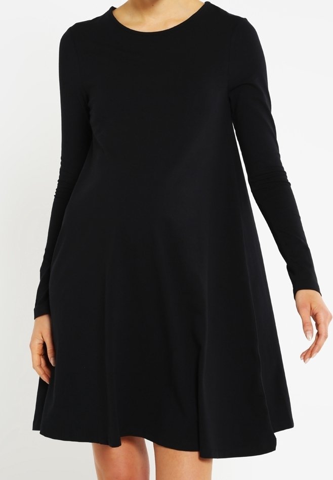 Femme portant une robe noire unie à manches longues et coupe évasée avec un col rond, debout devant un fond blanc.