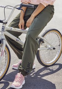 Groene joggers met taps toelopende boorden en zijnaad, gecombineerd met lichtroze sportschoenen. Een witte fiets met tan kleurige banden is zichtbaar.