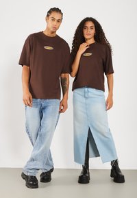 T-shirts oversize marron avec un logo, associés à une jupe en denim bleu clair avec une fente. Portés avec des bottes compensées noires.