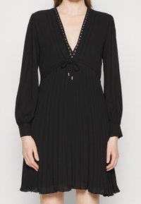 Abito nero plissettato con maniche lunghe, scollo a V adornato con borchie metalliche, vita con cordoncino e tessuto morbido e fluido.