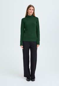 Groene, getailleerde turtleneck trui gemaakt van een textuurmateriaal, gecombineerd met zwarte wijde broek en zwarte schoenen.