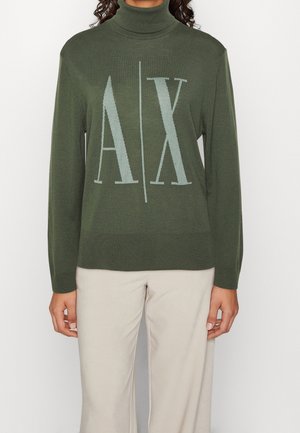 Person trägt einen olivgrünen Rollkragenpullover mit großen "A | X" Buchstaben auf der Vorderseite und hellbeige Hosen vor weißem Hintergrund.