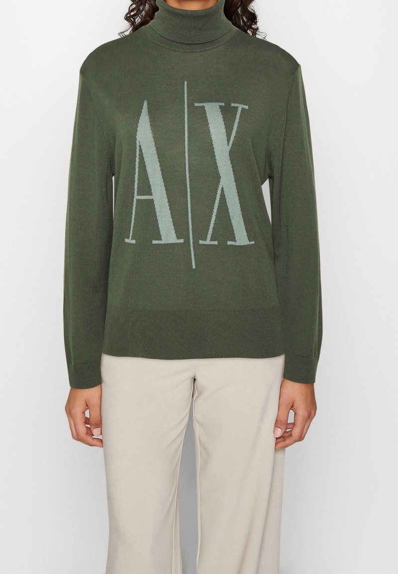 Personne portant un pull à col roulé vert olive avec de grandes lettres "A | X" sur le devant et un pantalon beige clair sur fond blanc.