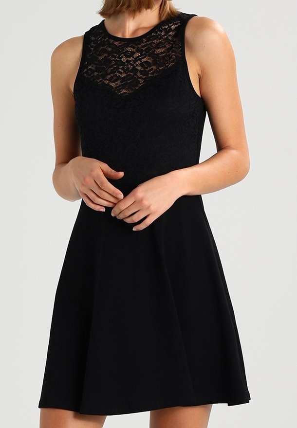 Femme portant une robe noire sans manches avec un détail en dentelle sur le haut de la poitrine et une taille ajustée, debout, les mains légèrement jointes devant elle.