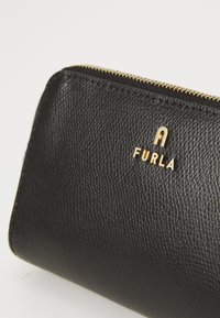 Portafoglio in pelle nera texturizzata con zip dorata e piccolo logo Furla dorato sul fronte, su sfondo bianco.