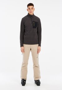 Zwart en grijs langemouwpullover met ritssluiting in de hals, voorzien van een borstzak. Beige sneeuwbroek met ritssluitingen bij de boorden en zwarte skischoenen.