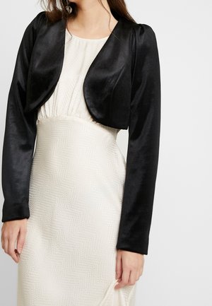 Femme portant une robe blanche ajustée à texture avec une taille haute et un boléro noir en satin à manches longues cropped.